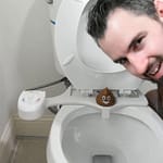 Man leaning over a toilet with a bidet attachment and a decorative item on the seat.