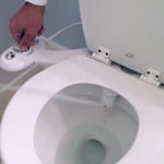 Hand operating the control panel of a bidet attachment on a toilet.