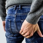 A man in a gray sweater holding his buttocks while wearing denim jeans.