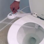 Hand operating the control panel of a bidet attachment on a toilet.