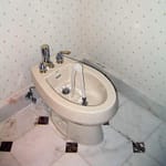 A white bidet with chrome fixtures and running water in a bathroom corner.