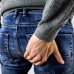 A man in a gray sweater holding his buttocks while wearing denim jeans.