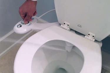 Hand operating the control panel of a bidet attachment on a toilet.
