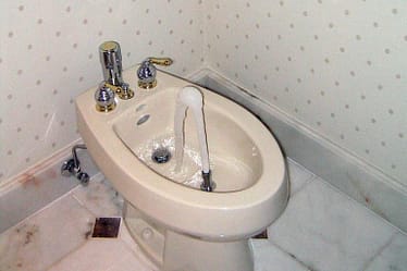 A white bidet with chrome fixtures and running water in a bathroom corner.