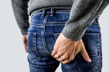 A man in a gray sweater holding his buttocks while wearing denim jeans.