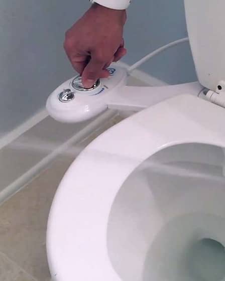 Hand operating the control panel of a bidet attachment on a toilet.
