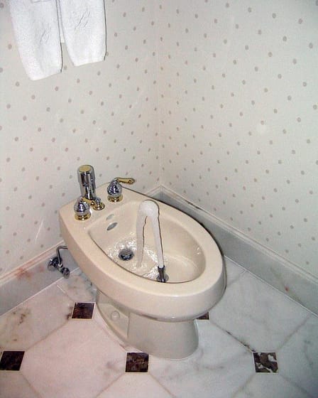 A white bidet with chrome fixtures and running water in a bathroom corner.