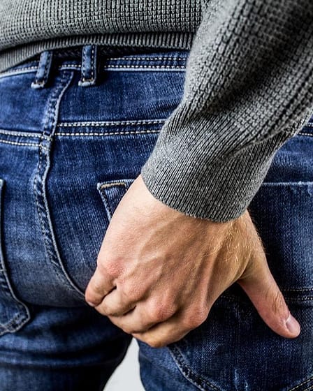 A man in a gray sweater holding his buttocks while wearing denim jeans.