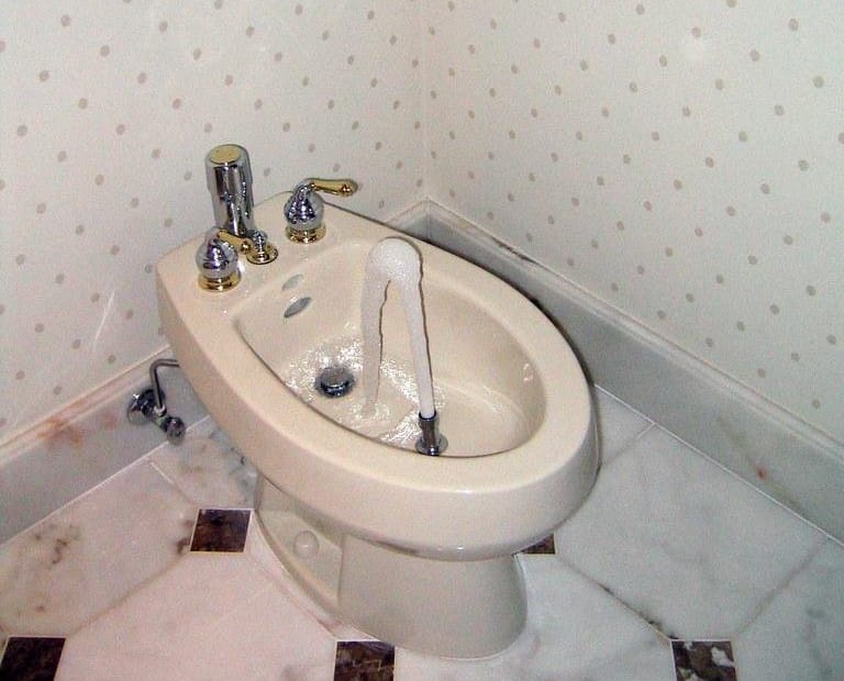 A white bidet with chrome fixtures and running water in a bathroom corner.