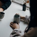 Two individuals holding coffee mugs on a table, with one mug being gray and the other white.