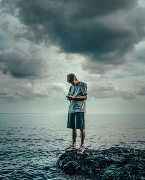 Man in a gray t-shirt and shorts standing on a rock near the sea under a cloudy sky.