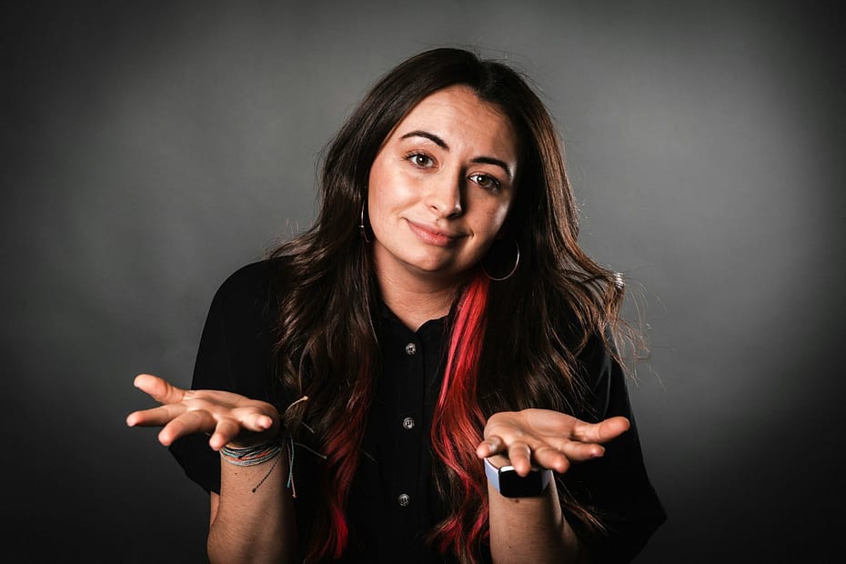 A woman with long hair featuring red highlights, wearing a black shirt and making a shrugging gesture.